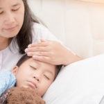 sick child laying in bed with mother measuring fever with her hand